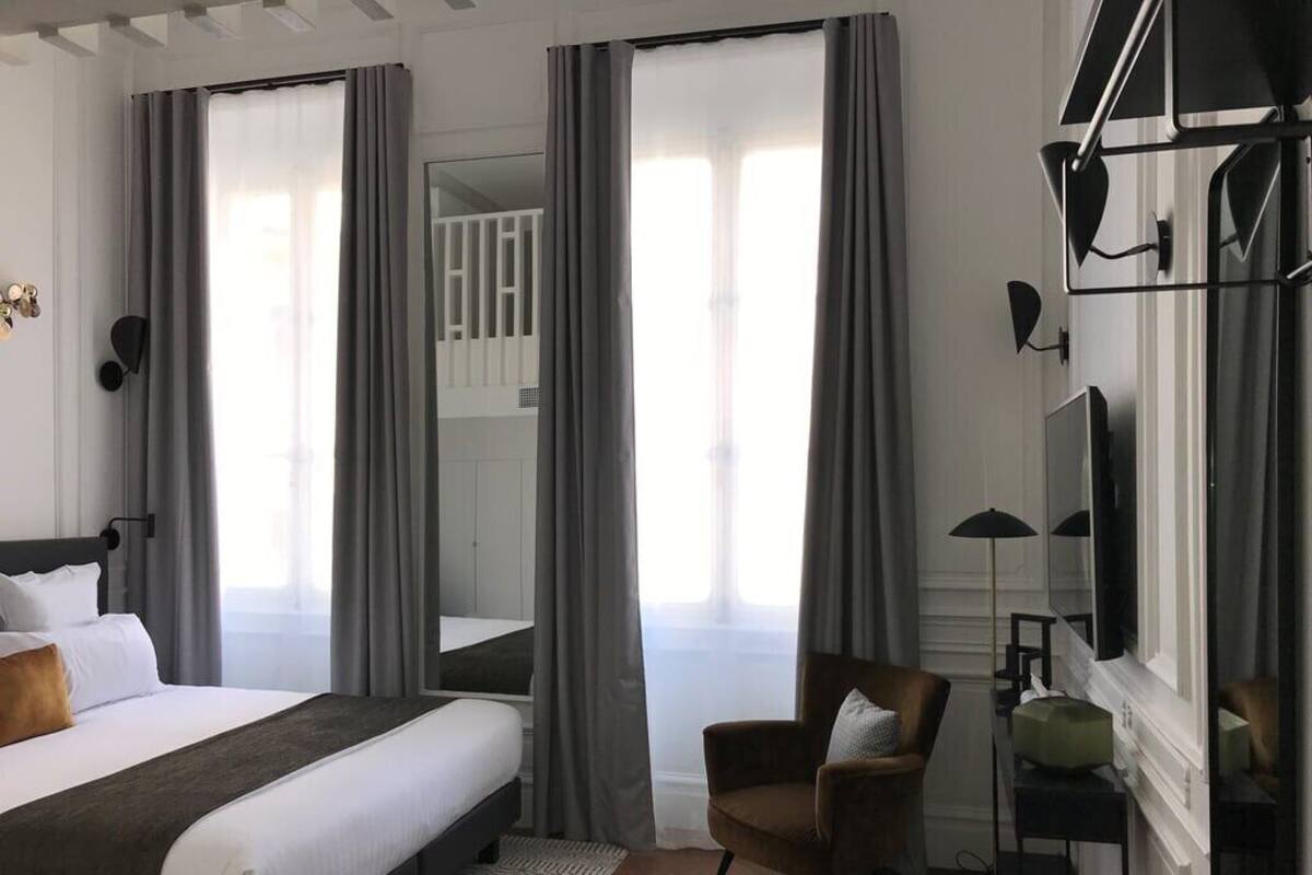 A hotel room with a bed, a chair, a desk, and a large window with curtains.