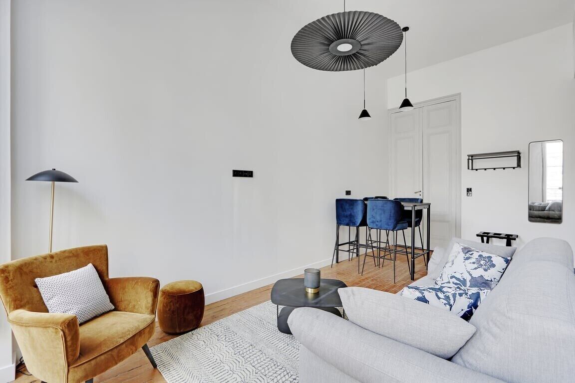 A modern living room with a sofa, armchair, and ottoman, a dining area with chairs and a table, and a large mirror on the wall.