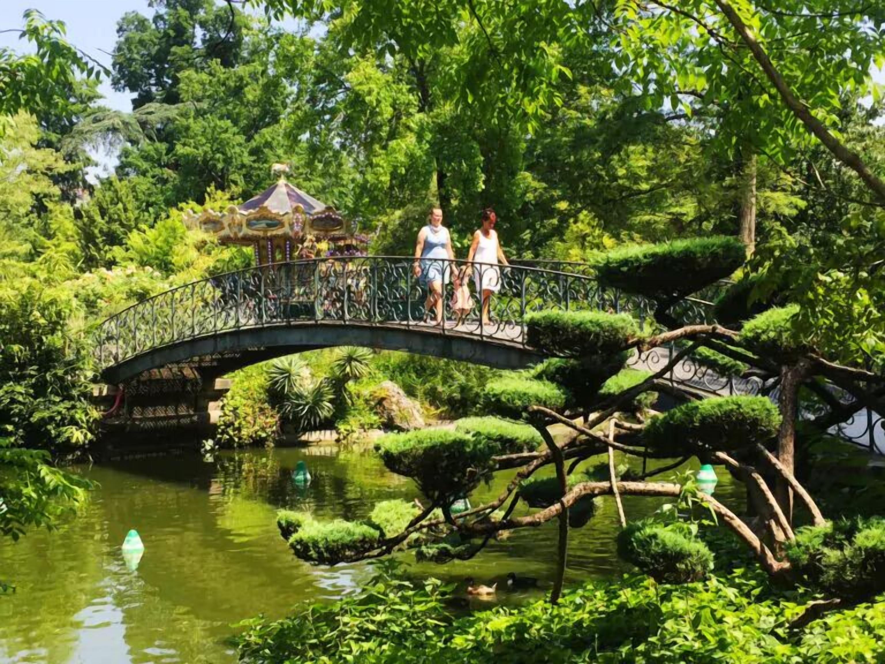 A bridge over a pond with lush greenery and a gazebo in the background.