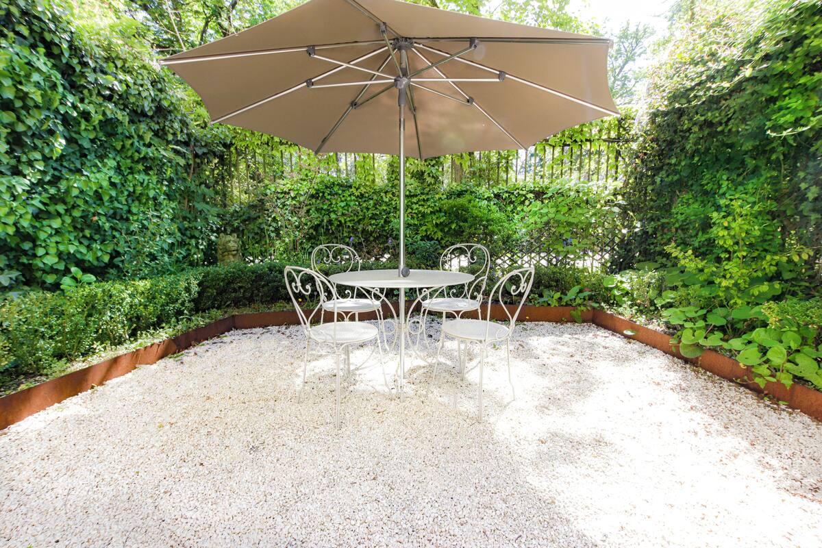 A round table with four chairs and a large umbrella in a garden setting.