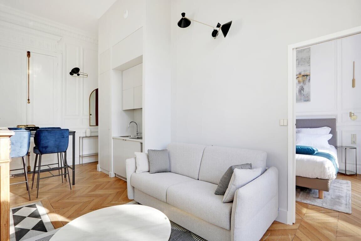 Un salón moderno y bien iluminado con un sofá, una mesa de comedor con sillas, una zona de cocina y un dormitorio con cama.