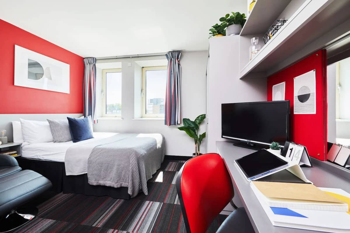 A modern hotel room with a bed, a desk with a computer, a TV, and a red chair.