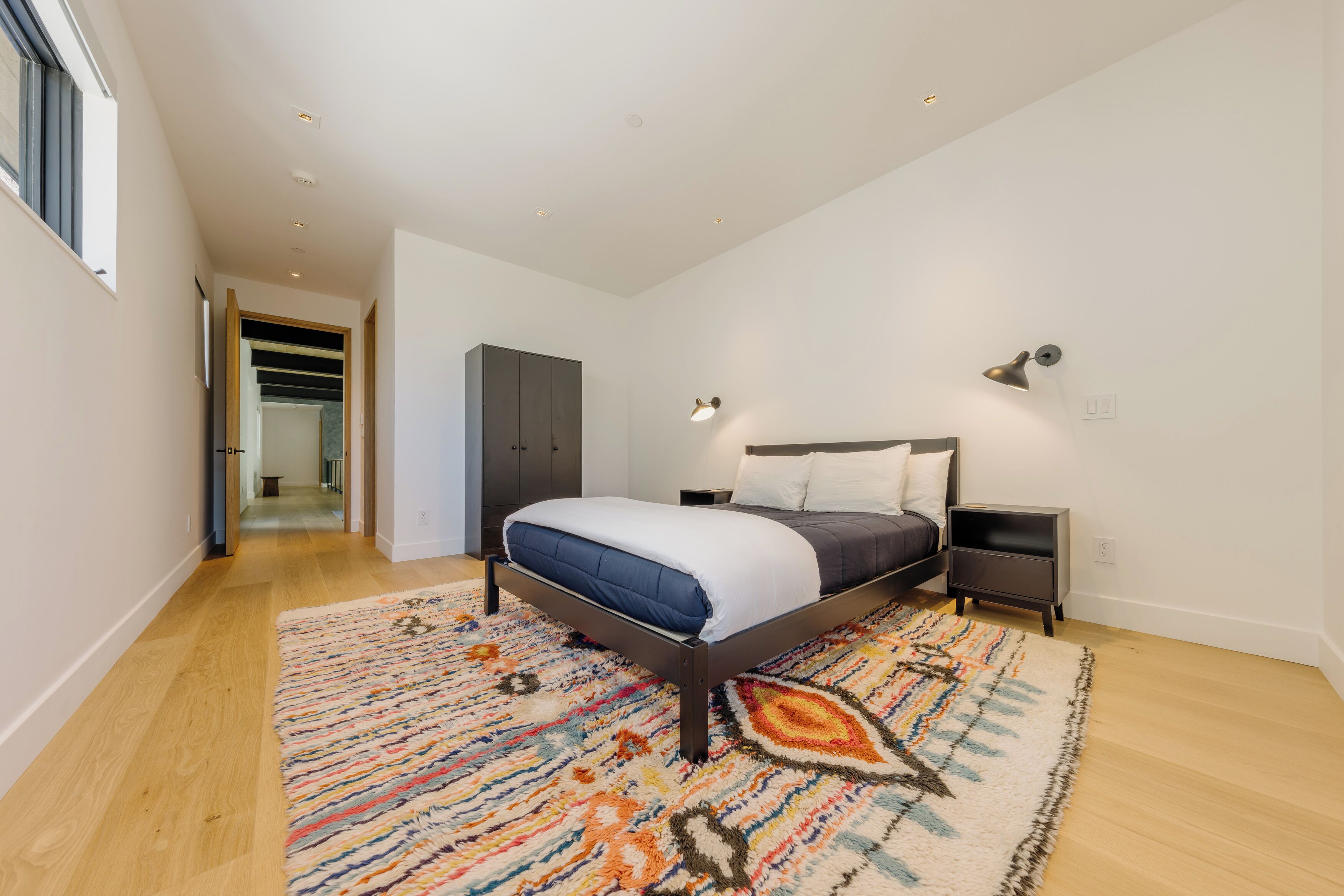 Ein Schlafzimmer mit einem Bett, einem Nachttisch, einer Lampe, einem gemusterten Teppich und einem Flur, der zu einem anderen Raum führt.