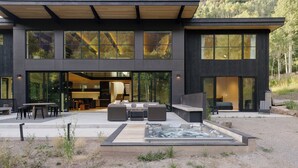 A modern two-story house with large windows, a patio with outdoor furniture, and a hot tub.