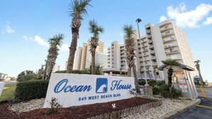 A sign for Ocean House at 949 West Beach Blvd. with palm trees and a multi-story building in the background. - Ocean House 1403 by Liquid Life (Gulf Shores)