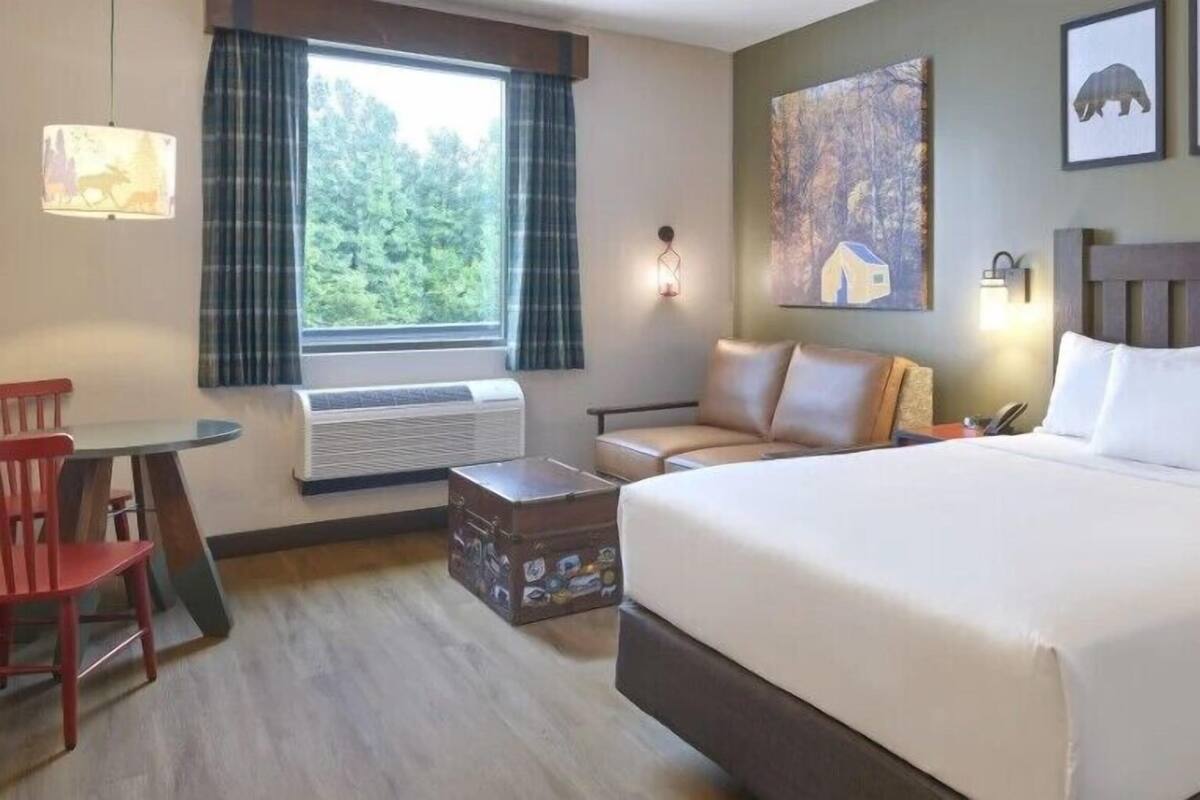 A hotel room with a bed, a desk, a chair, a window with curtains, and a wall-mounted lamp.