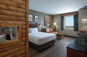 Una habitación de hotel con una cama, un sillón de cuero, una mesita, una lámpara y un ventanal con cortinas.