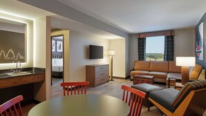 Une chambre d’hôtel avec une table à manger, des chaises rouges, un canapé, un téléviseur, une lampe et une vue sur l’extérieur.