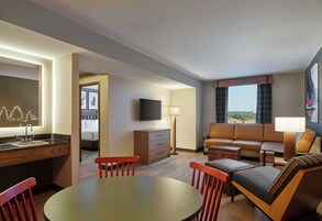 Habitación de hotel con una mesa de comedor, sillas rojas, un sofá, televisión, lámpara y vistas al exterior.