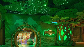 Un espacio interior temático con un arco en forma de árbol que conduce a una zona comercial, un letrero luminoso y un árbol con el tronco iluminado.