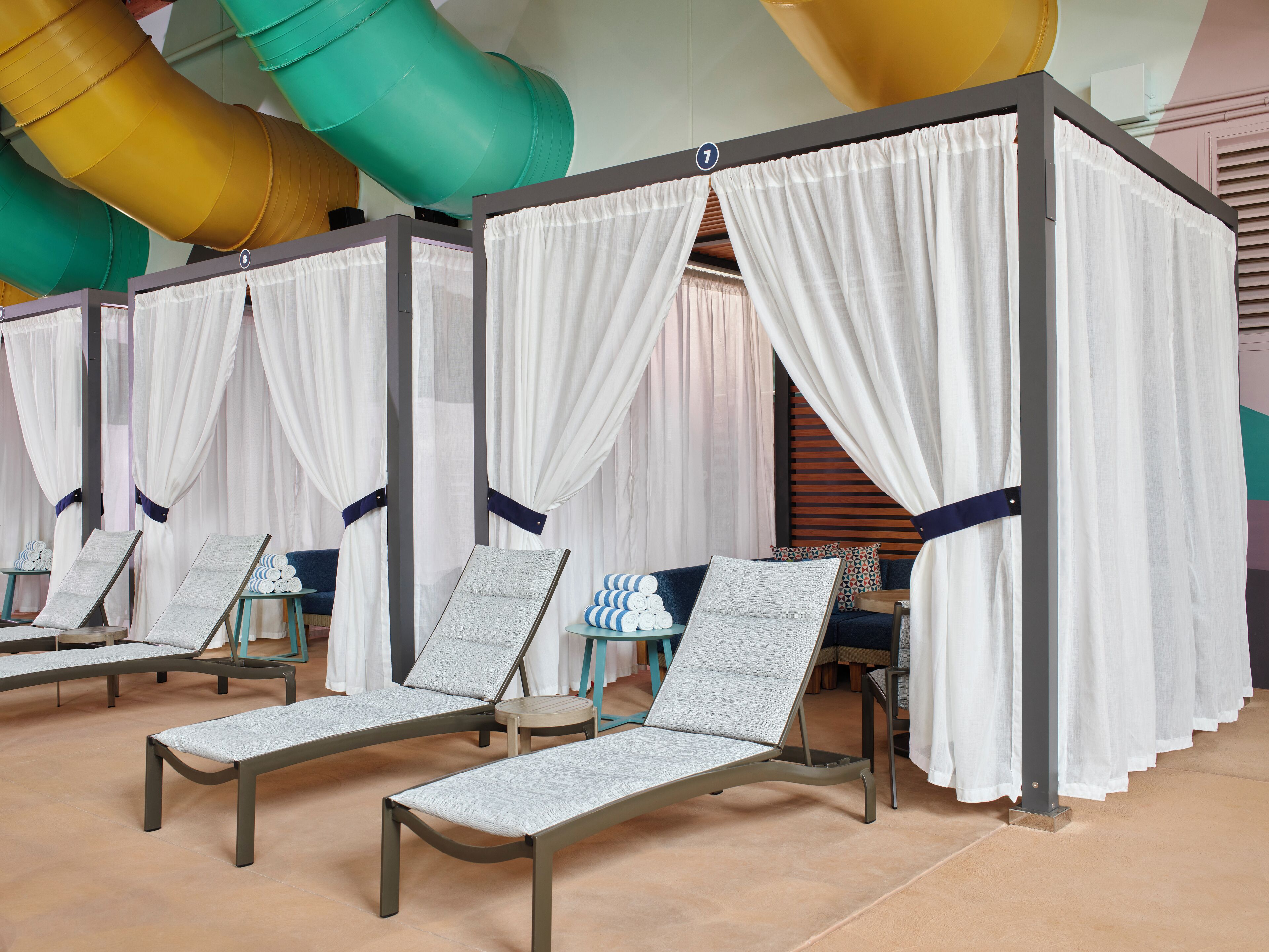 A row of sun loungers with white canopies, a pool with colorful slides, and a tiled floor.