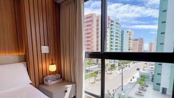 A hotel room with a bed, a bedside table with a lamp, and a view of a cityscape through large windows.