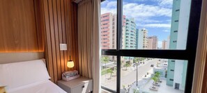 Habitación de hotel con cama, mesita de noche con lámpara y vistas a un paisaje urbano a través de grandes ventanales.