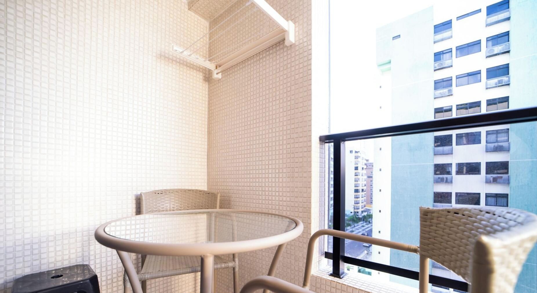Un balcón con una mesa redonda y dos sillas de mimbre, un muro de azulejos y vistas a edificios a través de una ventana.