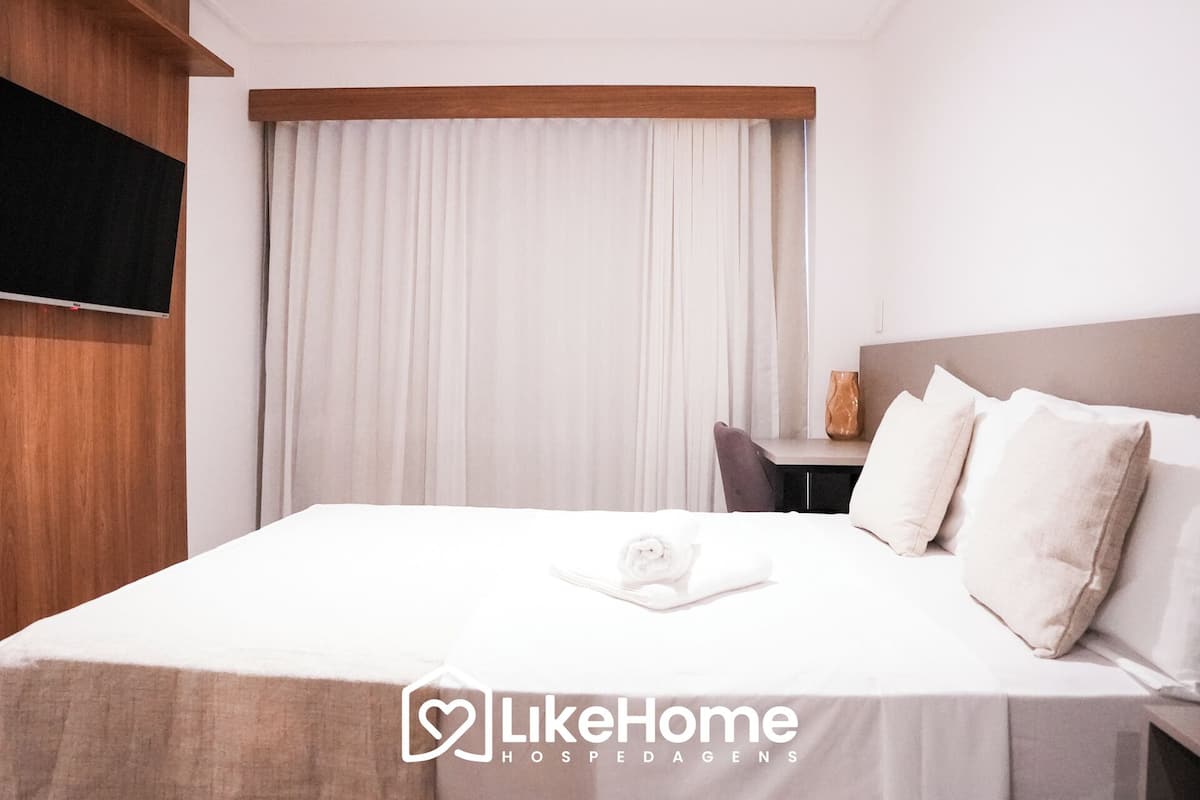 A hotel room with a neatly made bed, a wooden headboard, a small table with a vase, and a large window with curtains.