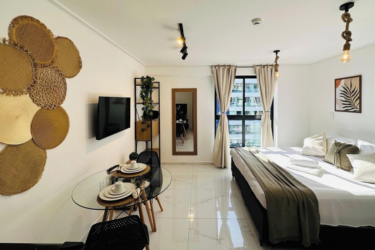 Habitación de hotel con cama, televisión, zona de comedor con mesa y sillas, y vista de la ciudad a través de la ventana.