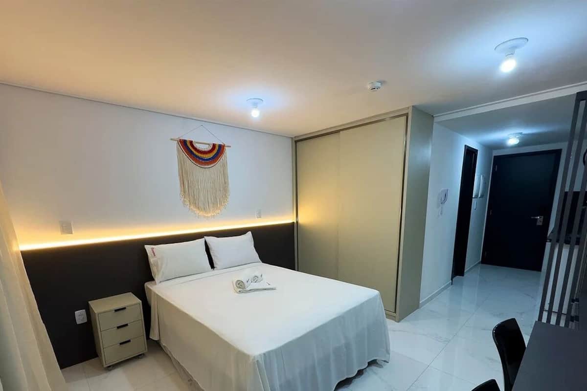 A modern hotel room with a neatly made bed, a bedside table, a small drawer unit, and a wall-mounted decorative piece.