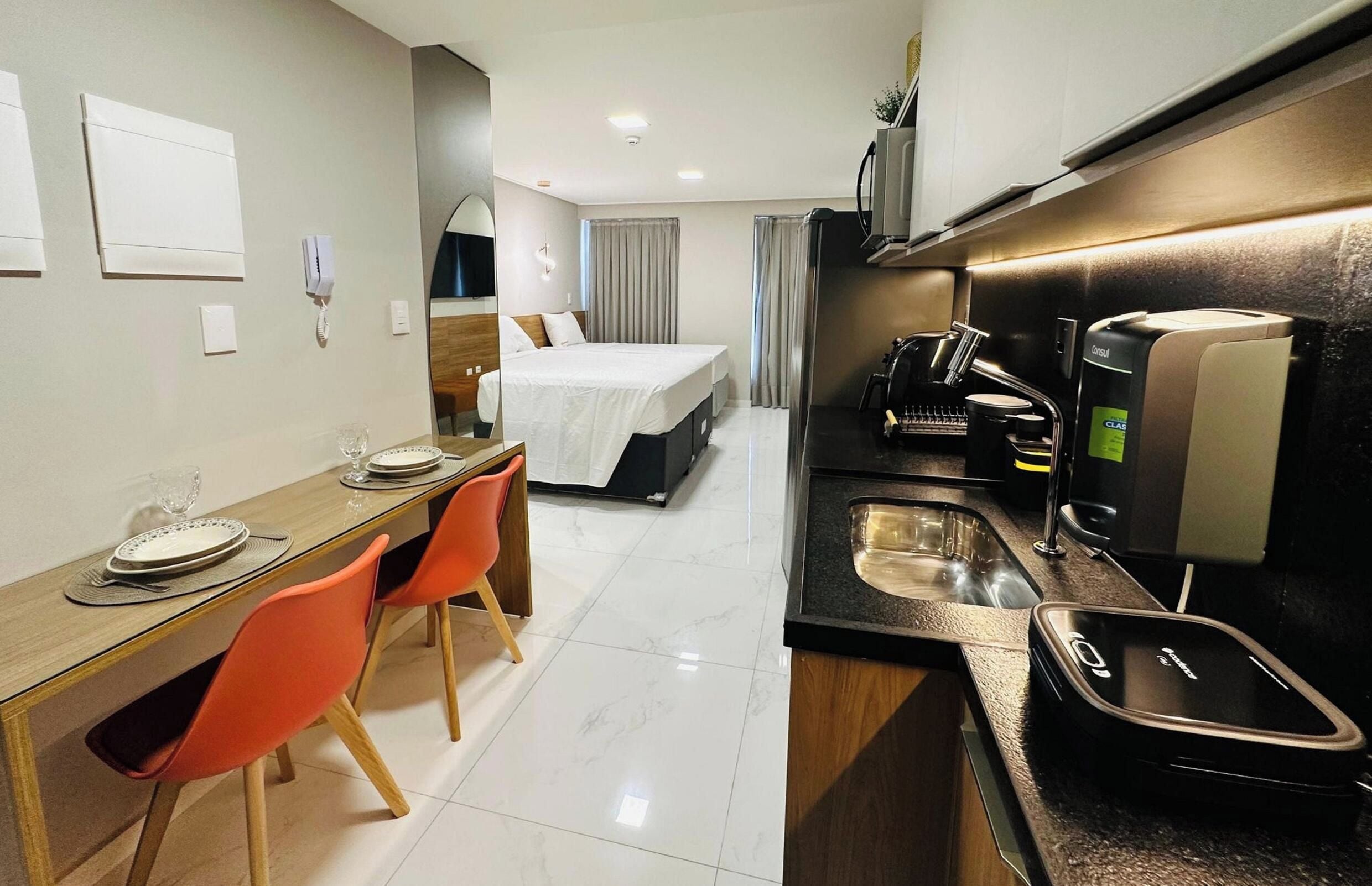 Una camera d'albergo compatta con un letto, un piccolo tavolo da pranzo con sedie, un angolo cottura con macchina del caffè e un bagno con lavandino.
