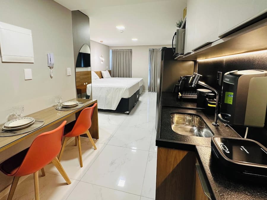 A compact hotel room with a bed, a small dining table with chairs, a kitchenette with a coffee machine, and a bathroom with a sink.