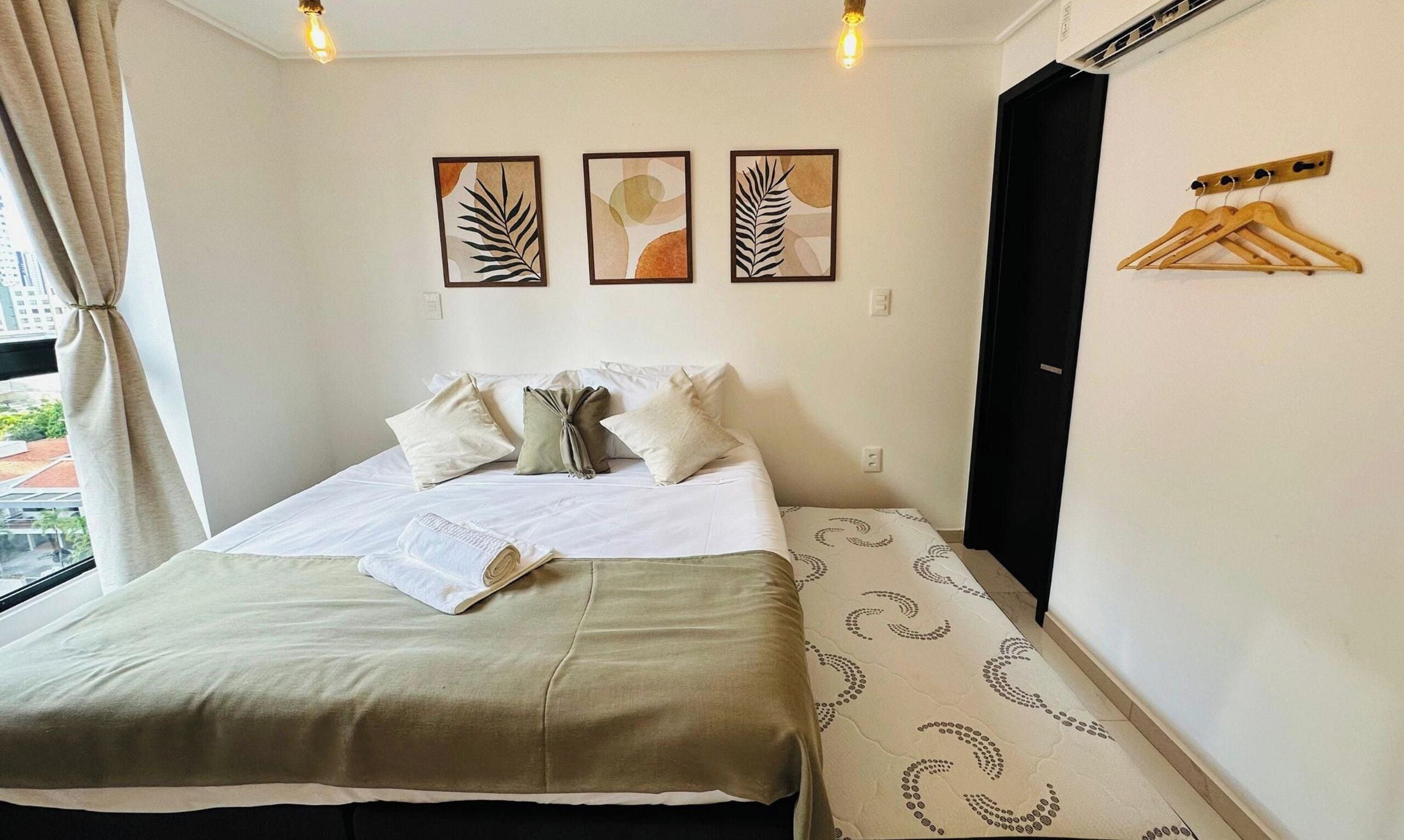 Una camera d'albergo con un letto, cuscini, una coperta, un appendiabiti a parete e tre opere d'arte incorniciate.