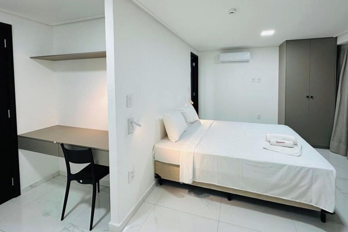 Un dormitorio moderno con cama, escritorio y silla. Se observa un armario y un equipo de aire acondicionado en la pared.