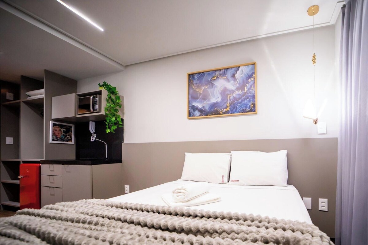 Habitación de hotel con una cama, un cuadro, una planta y una maleta roja.