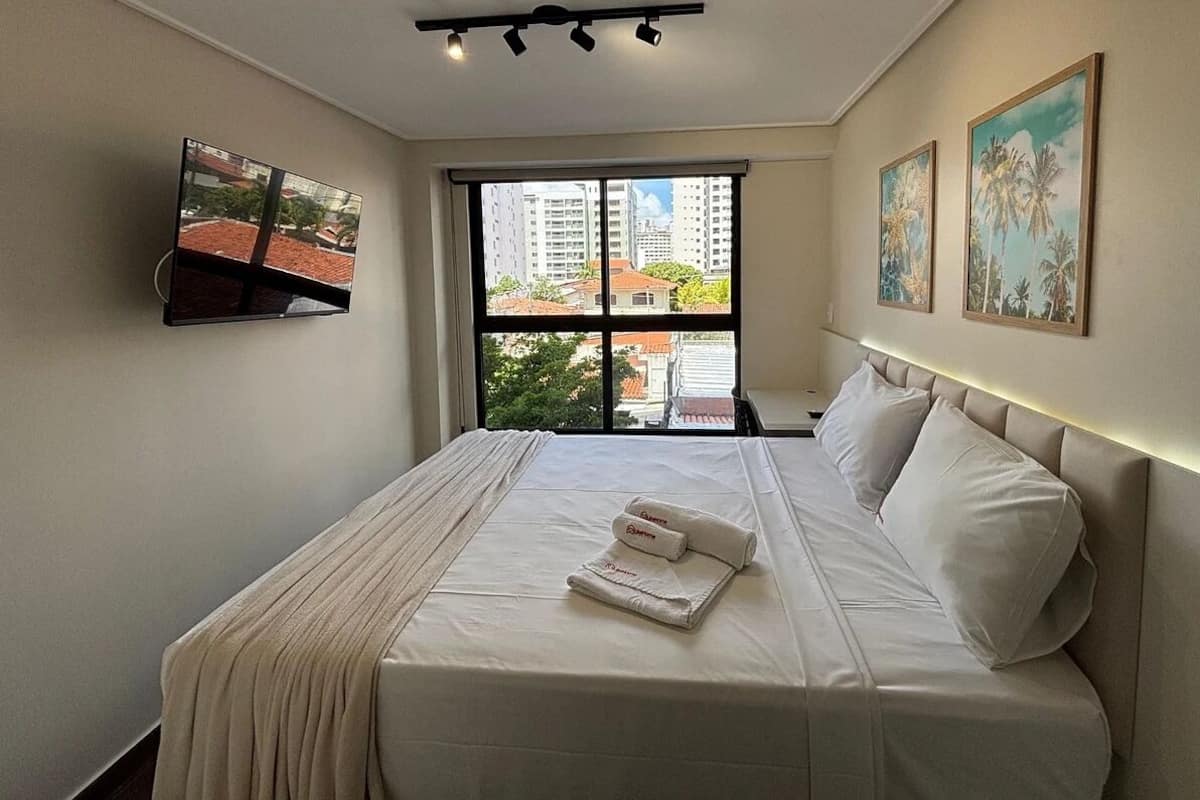 A hotel room with a bed, a flat-screen TV mounted on the wall, a large window with a city view, and two framed pictures on the wall.