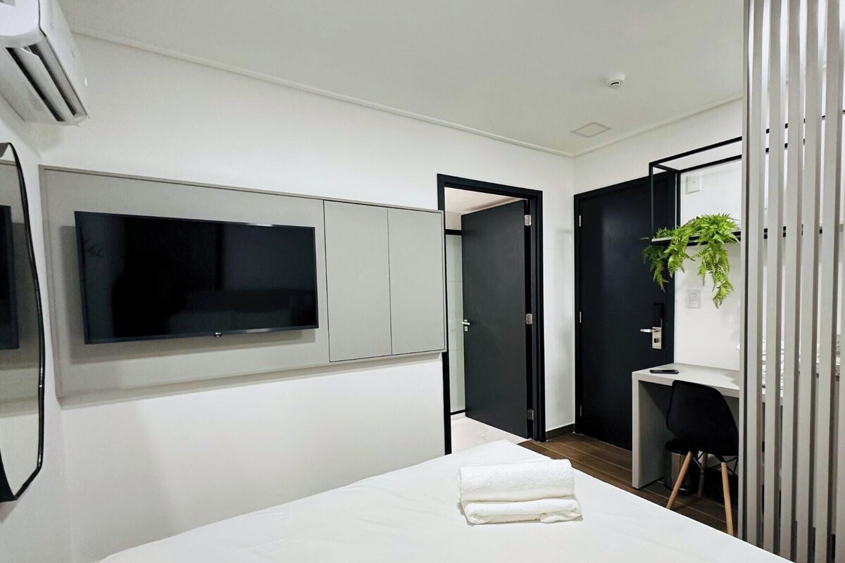 Un dormitorio moderno con cama, televisor, escritorio y planta.