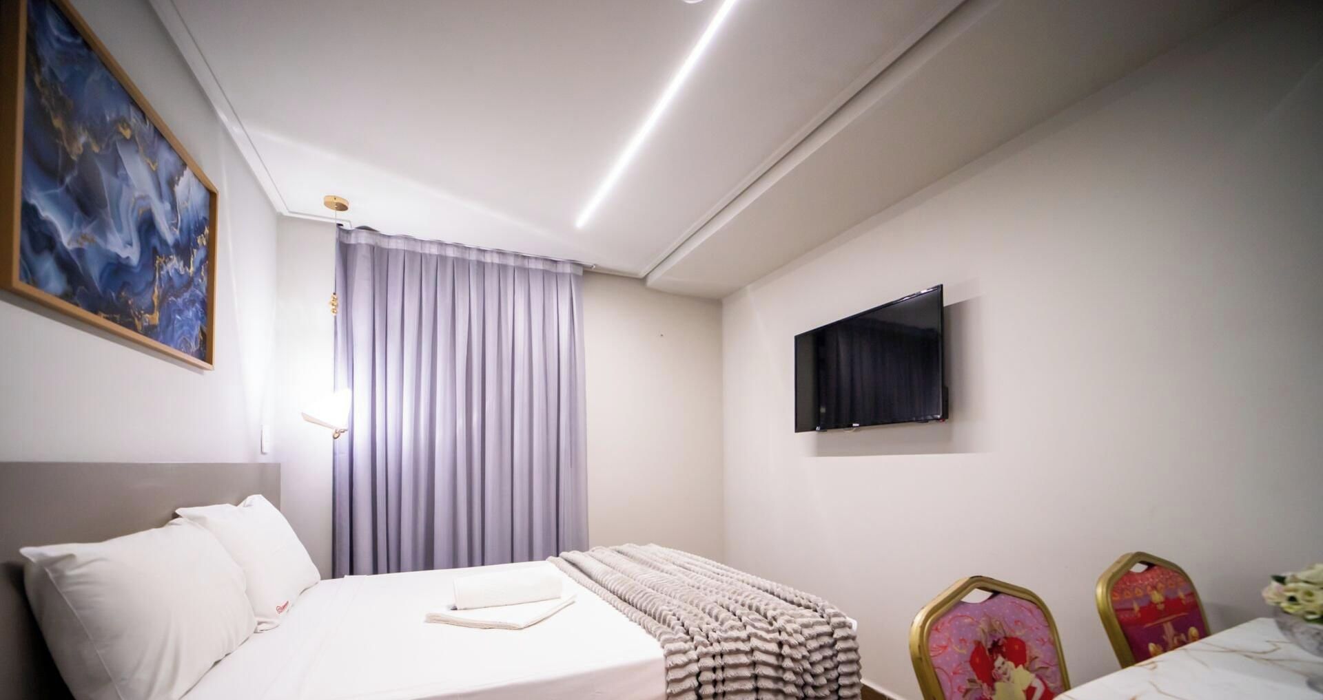 Una habitación de hotel con una cama, un televisor de pantalla plana en la pared, una ventana con cortinas y un cuadro en la pared.