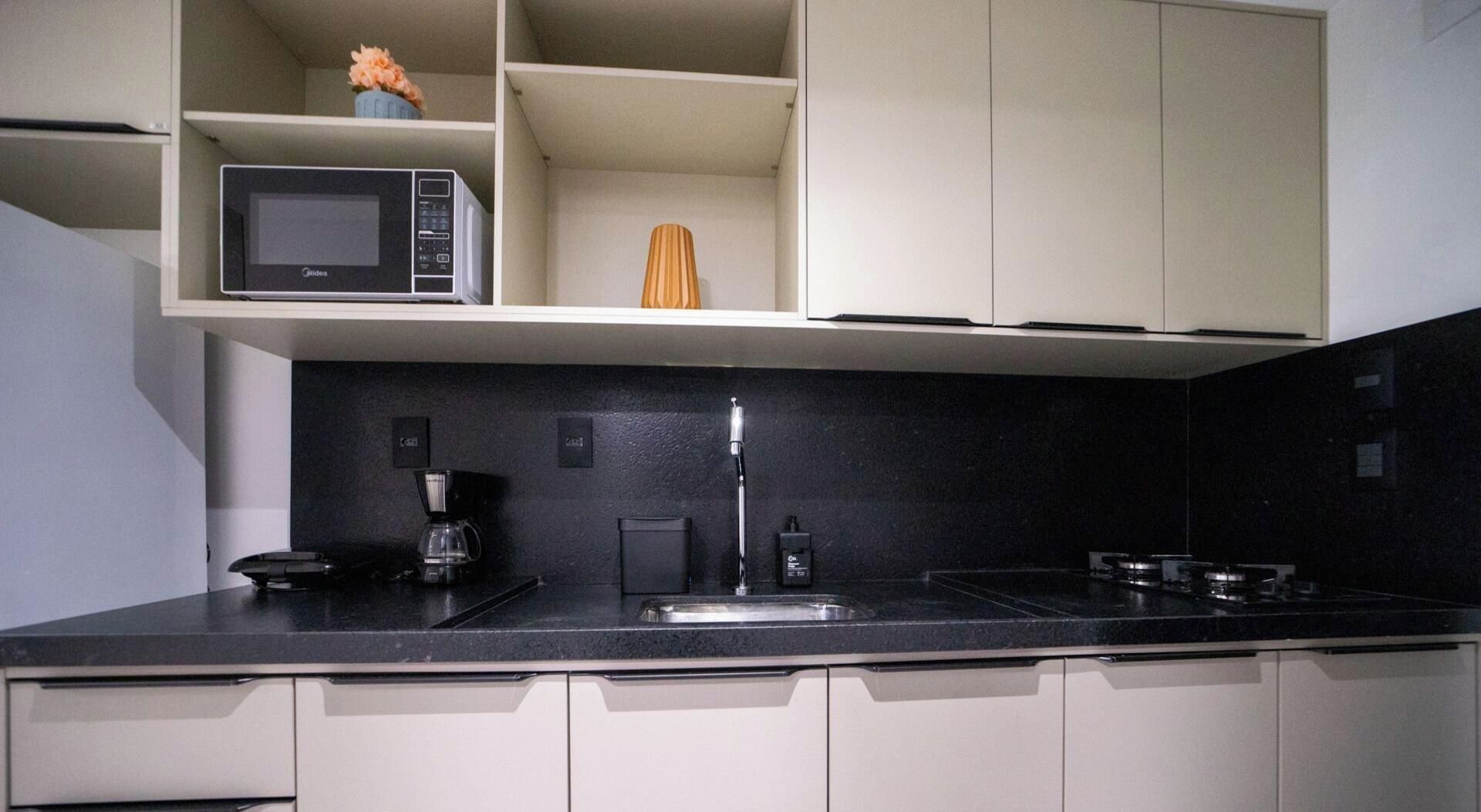 Una cocina moderna con encimera negra, microondas integrado, cafetera y lavamanos.