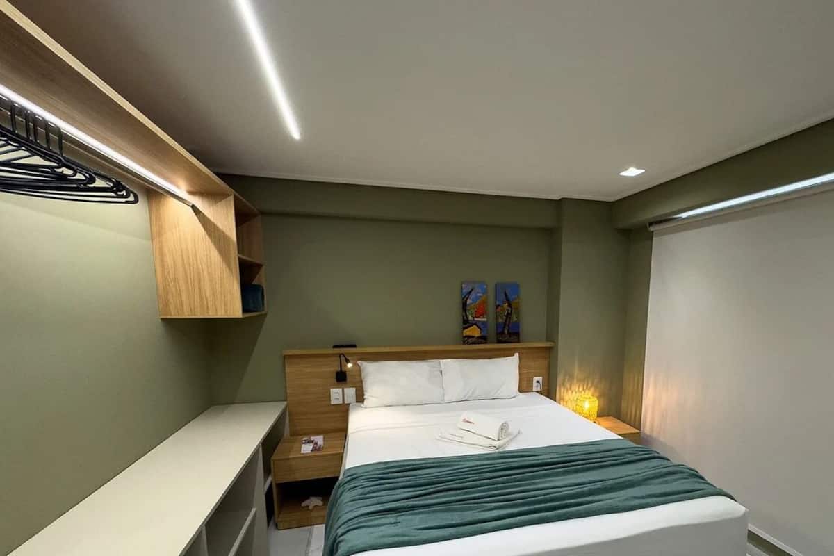 A modern hotel room with a neatly made bed, a bedside table, a built-in shelf, and a wall-mounted artwork.