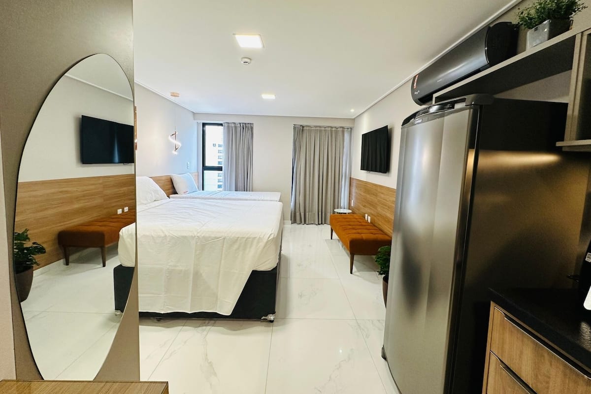 A modern hotel room with a large bed, a sofa, a dining table, and a kitchen area with a refrigerator.