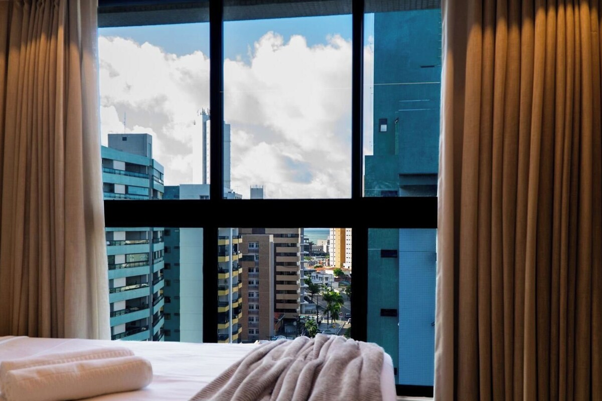 Una habitación de hotel con un ventanal grande, una cama con manta blanca y cortinas.