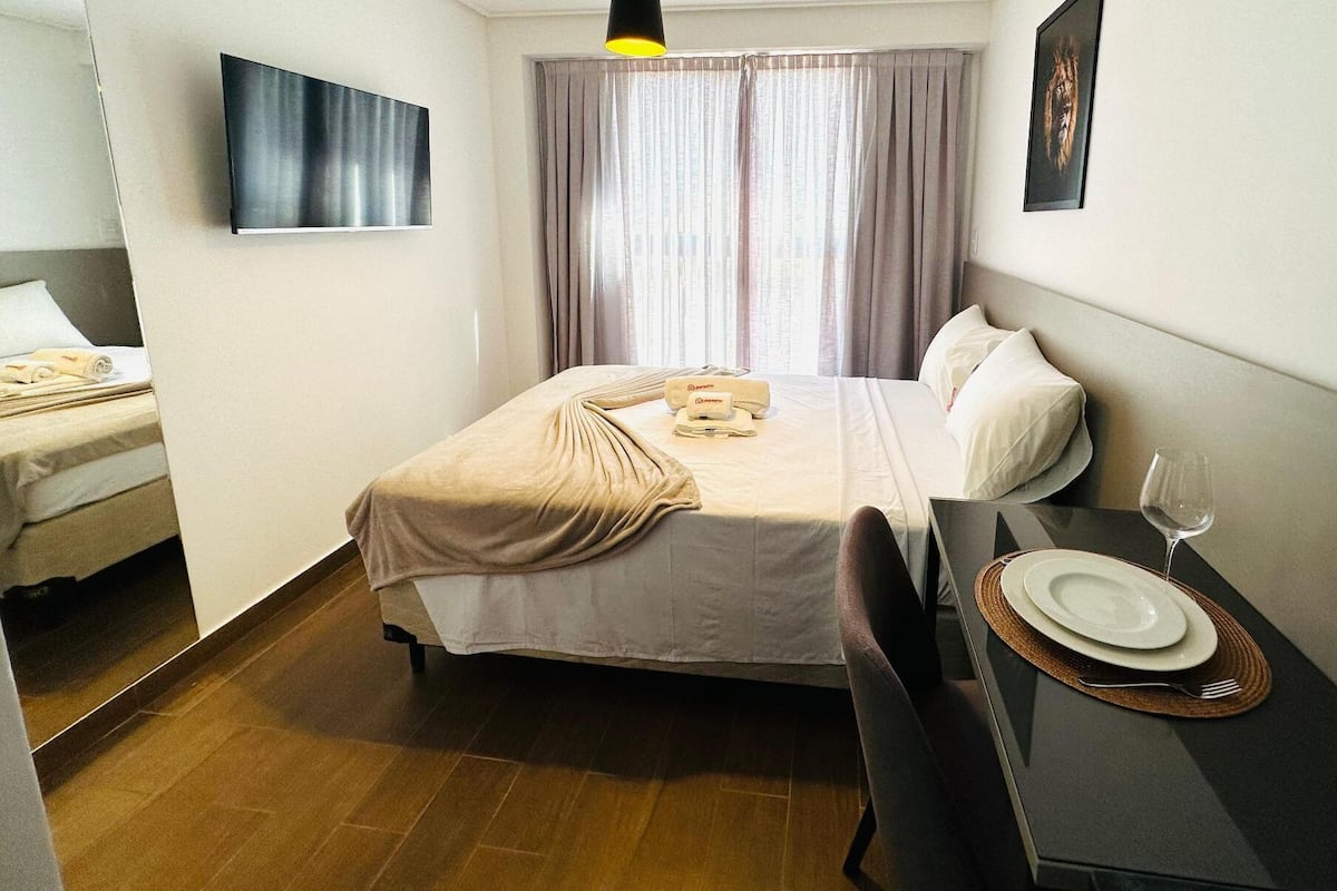 A hotel room with a bed, a bedside table with a glass of water, a framed picture on the wall, and a window with curtains.