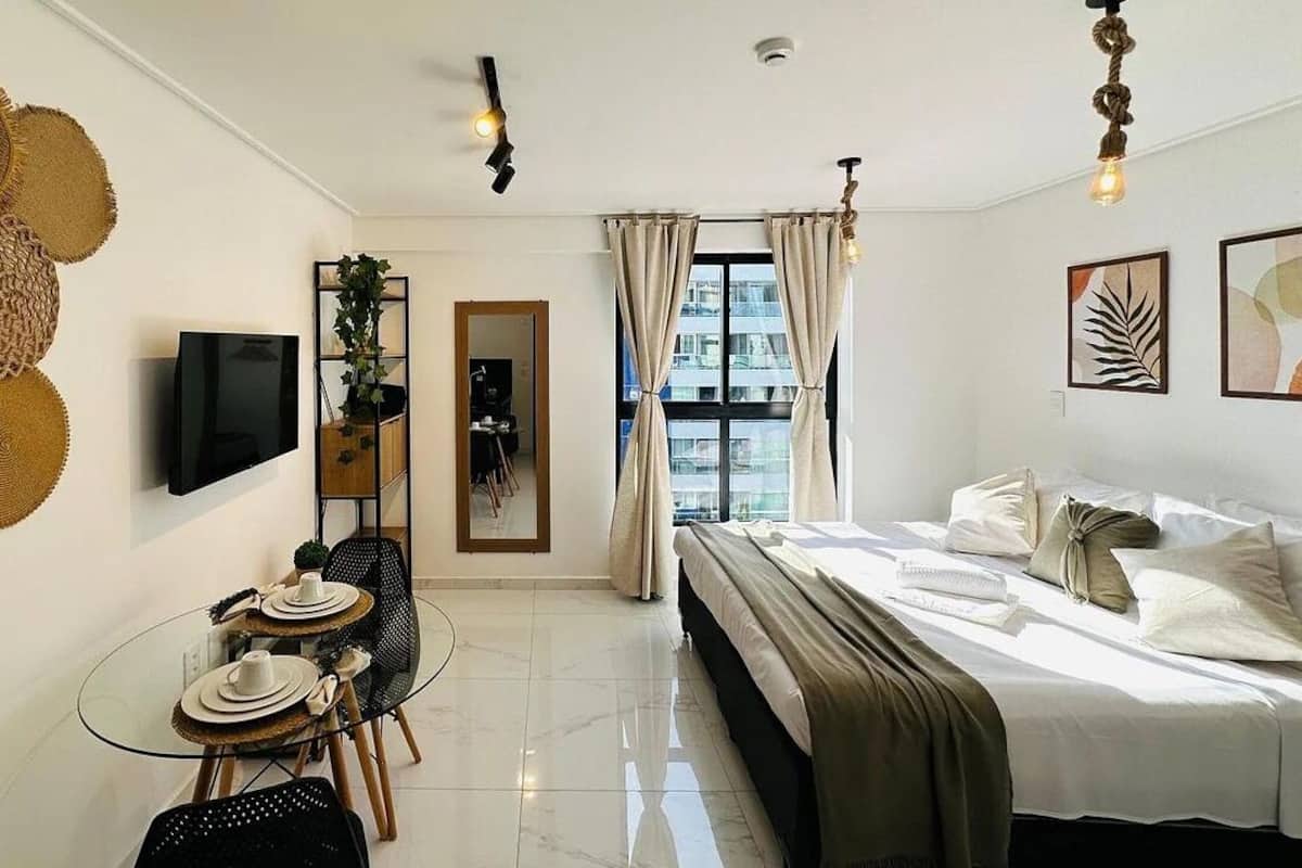 A hotel room with a bed, a TV, a dining table with chairs, and a large window with curtains.