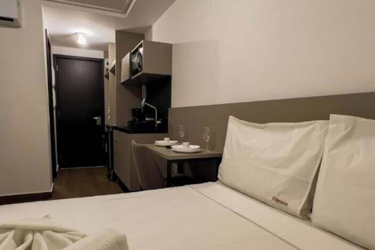 A hotel room with a bed, a small desk with a chair, a kitchenette, and a door leading to another room.