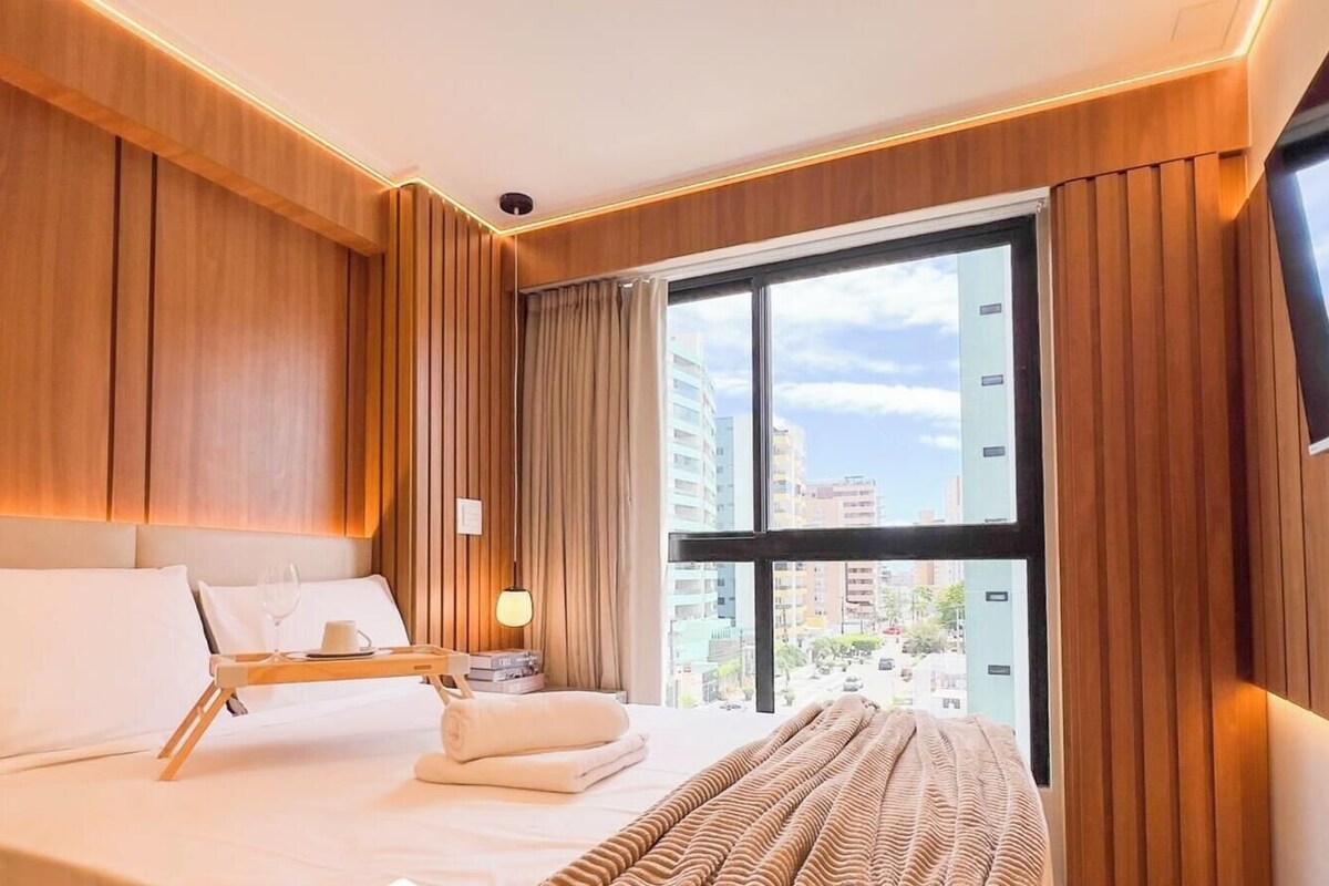 Una habitación de hotel con una cama, una mesita de noche con lámpara, una ventana con cortinas y vistas a la ciudad.