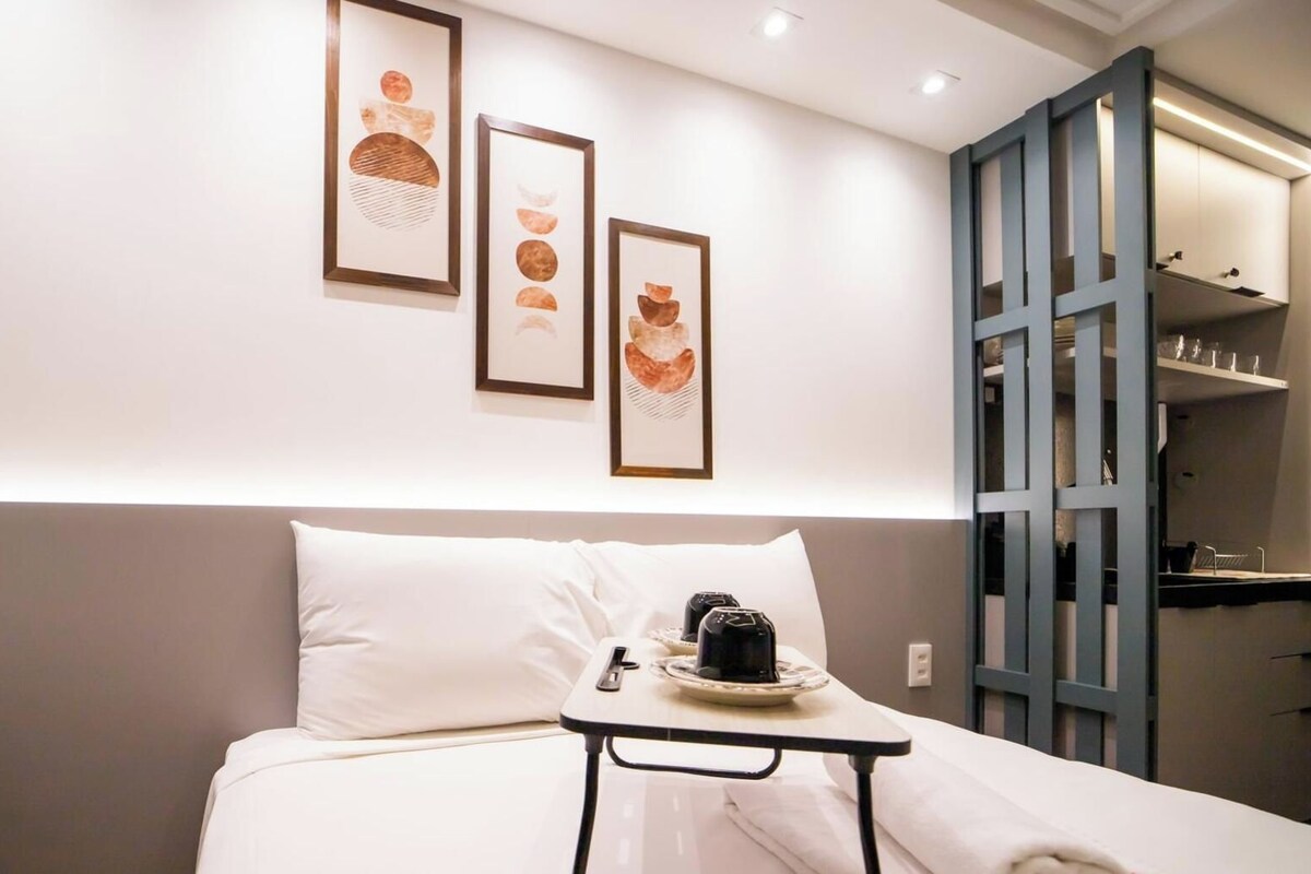 Habitación de hotel con cama, una mesita con una taza y un control remoto, y una obra de arte colgada en la pared.