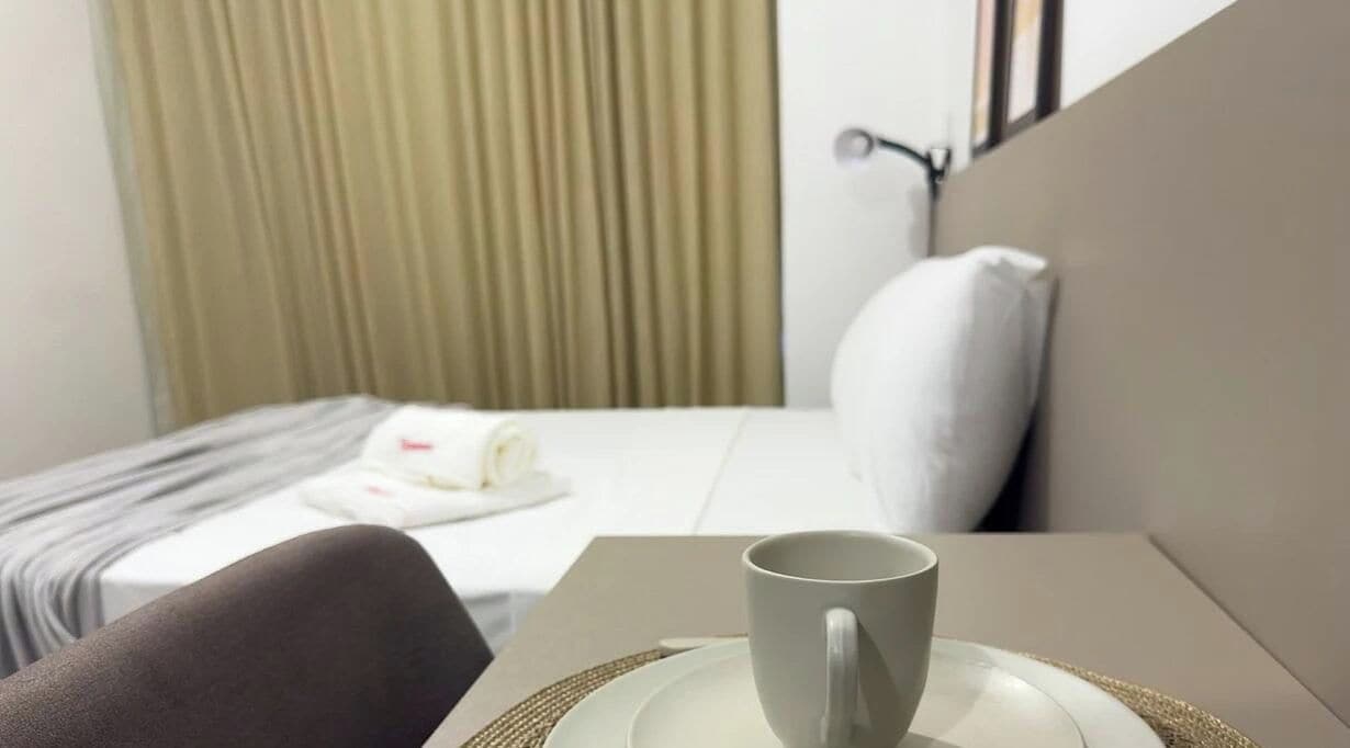A hotel room with a bed, a bedside table with a cup and saucer, a pillow, and a curtain.