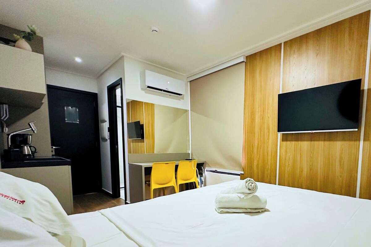 Habitación de hotel con cama, escritorio, silla, televisor y aire acondicionado de pared.