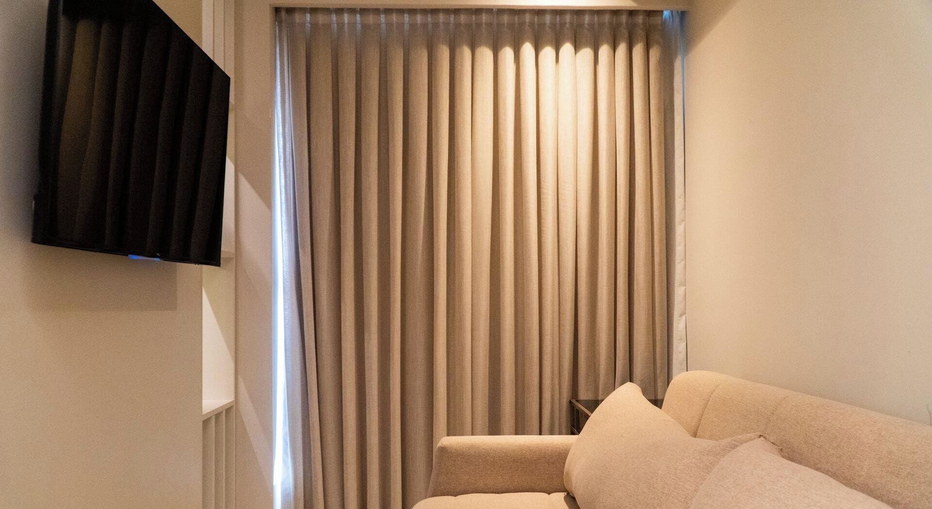 A living room with a sofa, a flat-screen TV mounted on the wall, and a window with vertical blinds.