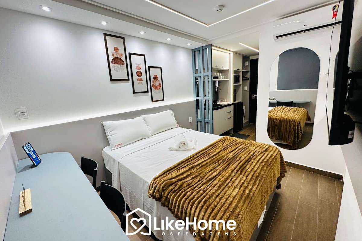 A modern hotel room with a bed, bedside table, and a desk. There are framed pictures on the wall and a mirror on the wall.