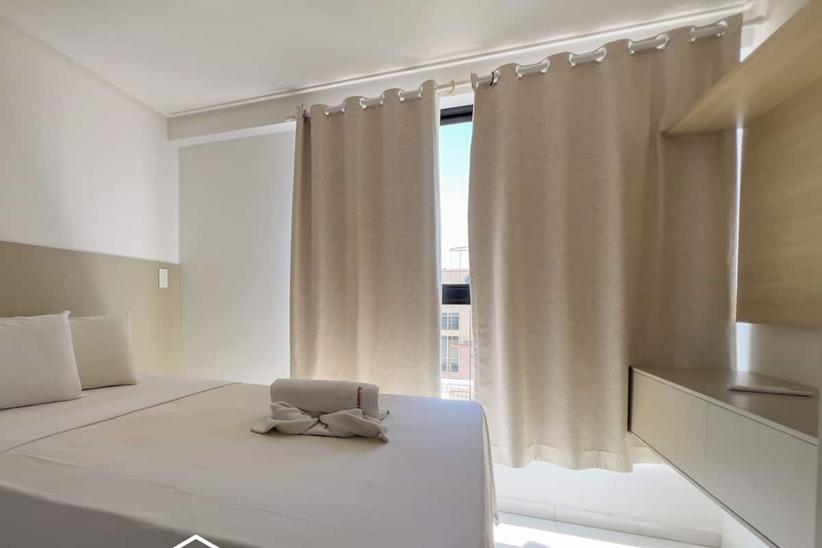 A hotel room with a bed, pillows, a folded towel, a window with curtains, and a mirror.