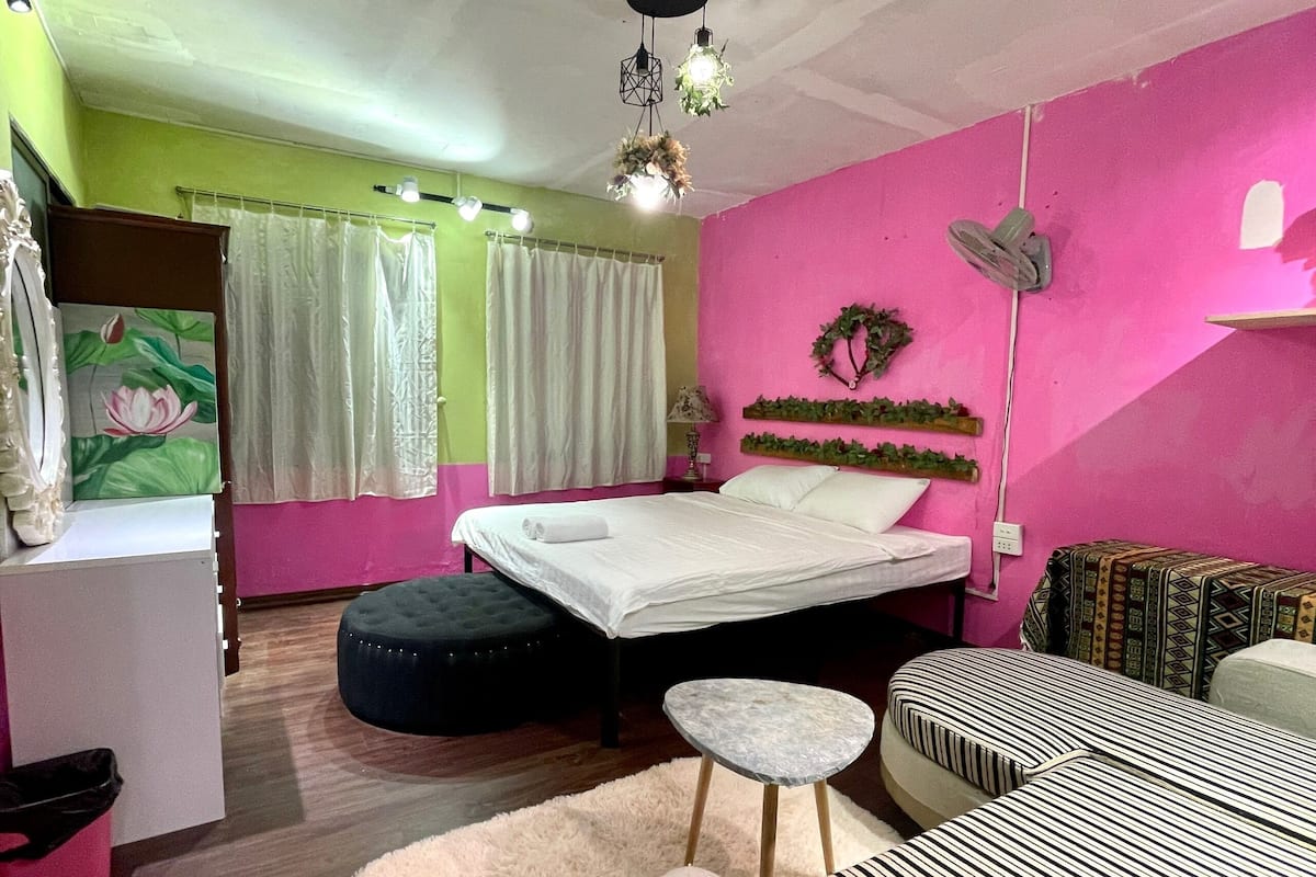 A bedroom with a bed, a small stool, a round ottoman, a fluffy rug, and a wall-mounted shelf with greenery.