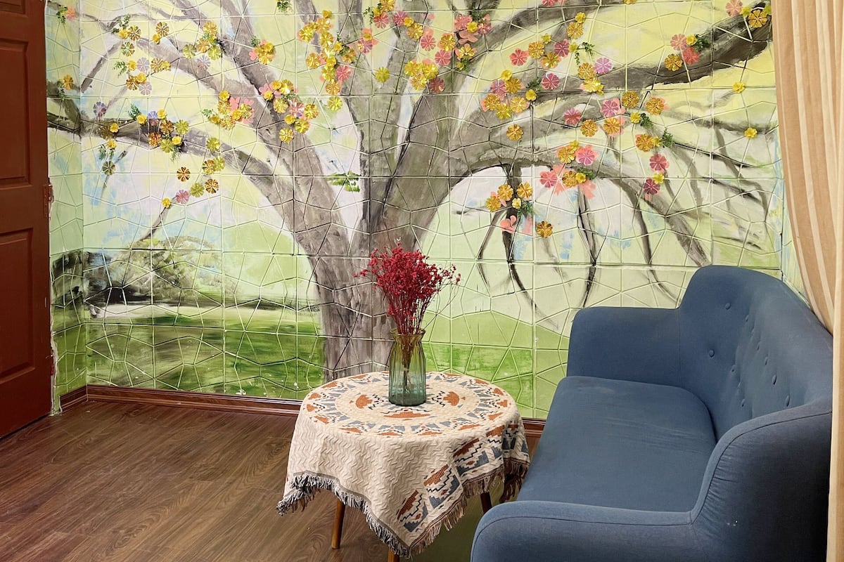 A room with a mural of a tree with flowers, a blue armchair, and a small table with a vase.