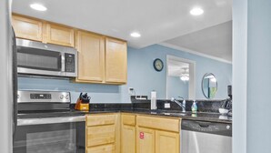 A modern kitchen with wooden cabinets, a microwave, and a dishwasher. There is a dining area with a table and chairs, and a view of a building outside.