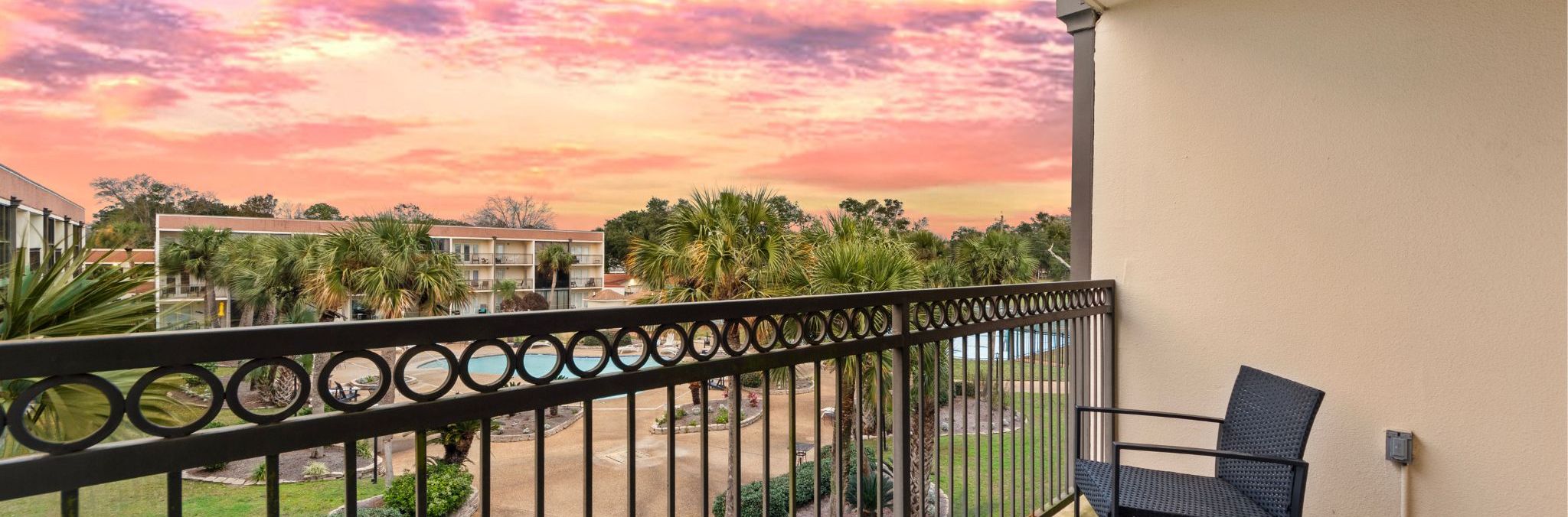 Een balkon met uitzicht op een complex van gebouwen, palmbomen en een lucht in de kleuren van de zonsondergang.