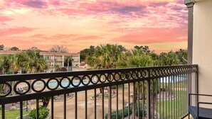 A balcony with a view of a building complex, palm trees, and a sunset sky.