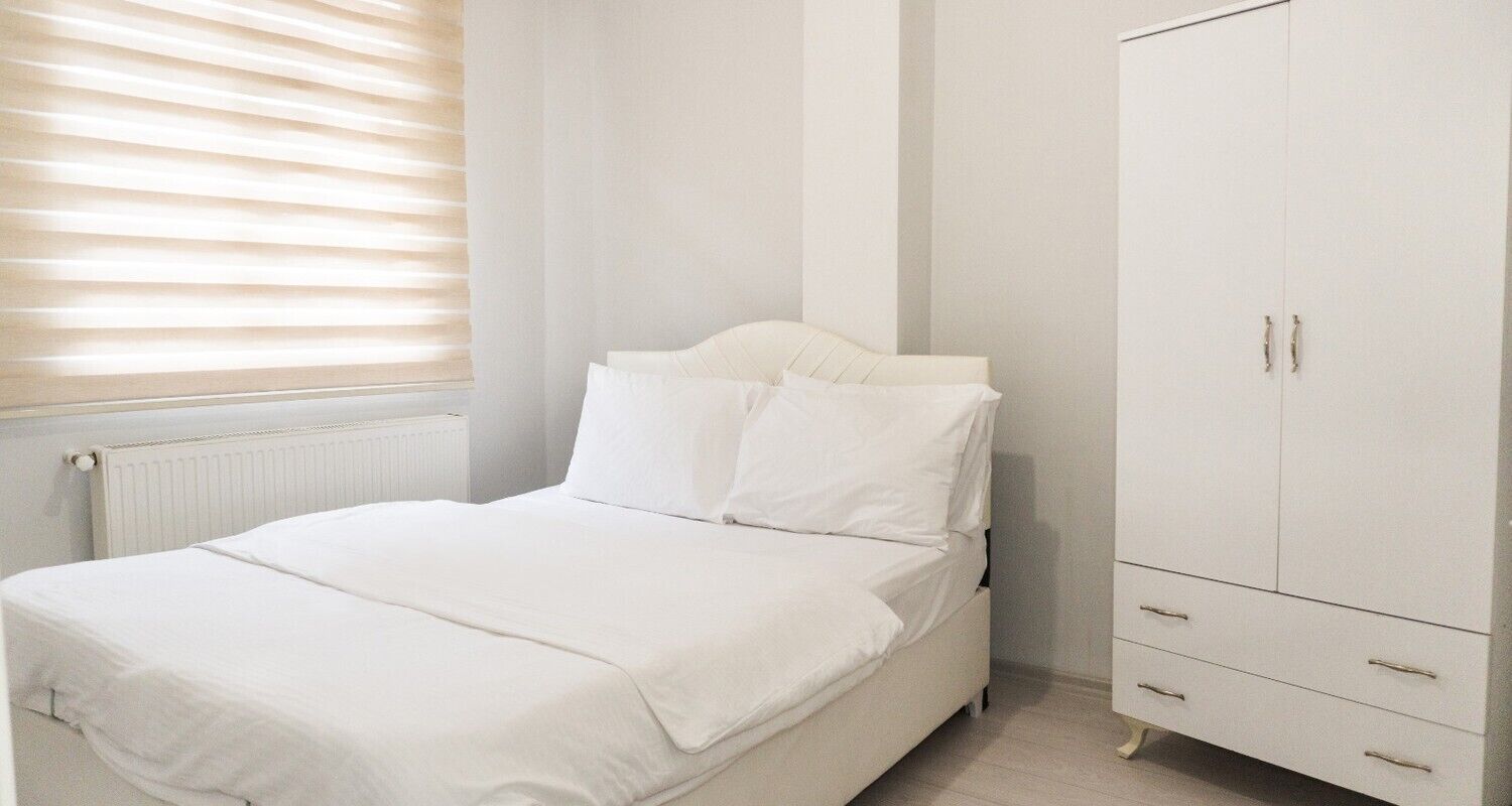 A bedroom with a bed, white bedding, a radiator, a window with blinds, and a white wardrobe with drawers.
