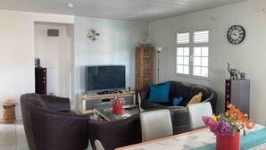 A modern living room with a flat-screen TV, dark sofas, a coffee table with a vase of flowers, and a dining area with a patterned tablecloth. - Villa Soleil Located in a quiet, serene residential area (Vauclin)